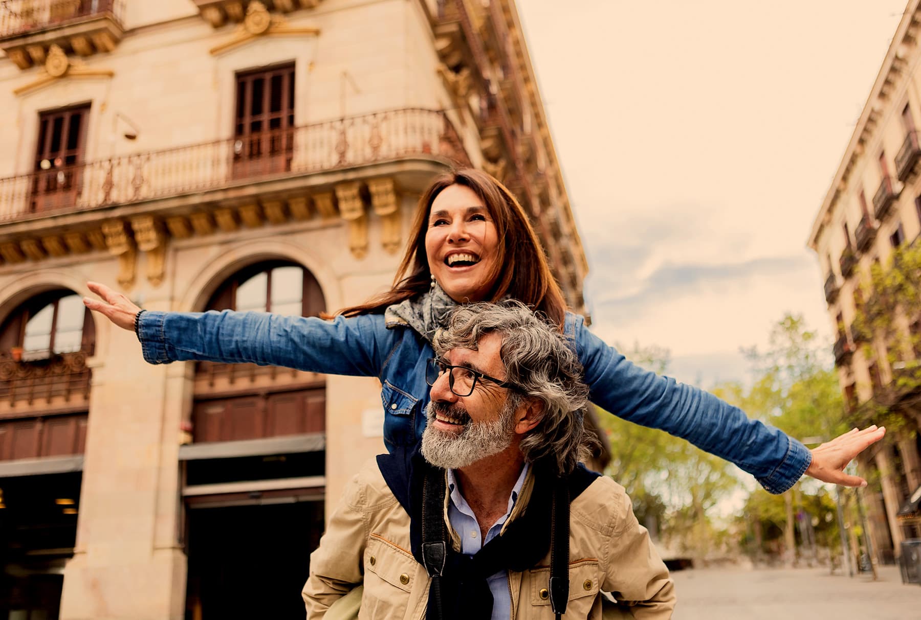 An older couple laughing in a European plaza, one carried on the other's back with both arms flung wide, a camera hanging on a strap at his chest