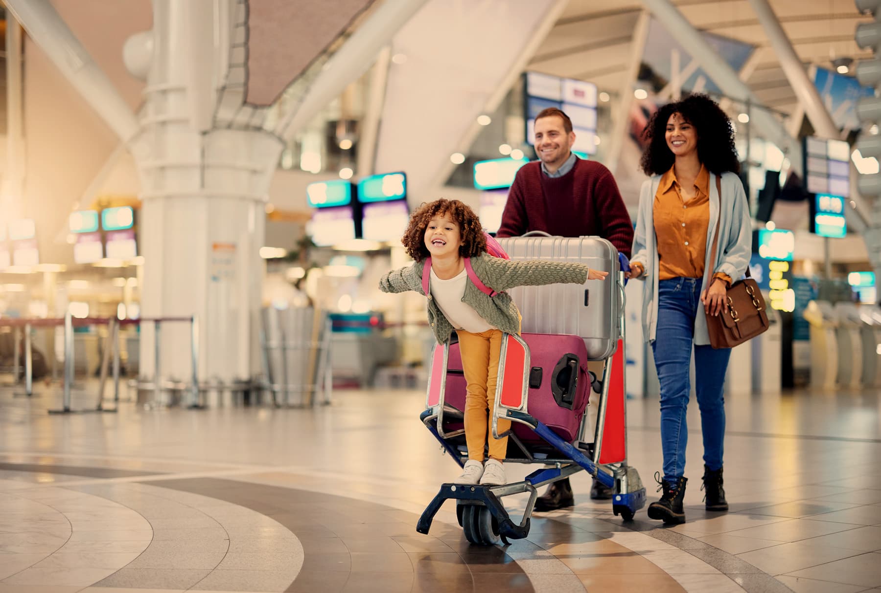 A family walking through an airport departures hall, a child riding the luggage trolley with arms outstretched