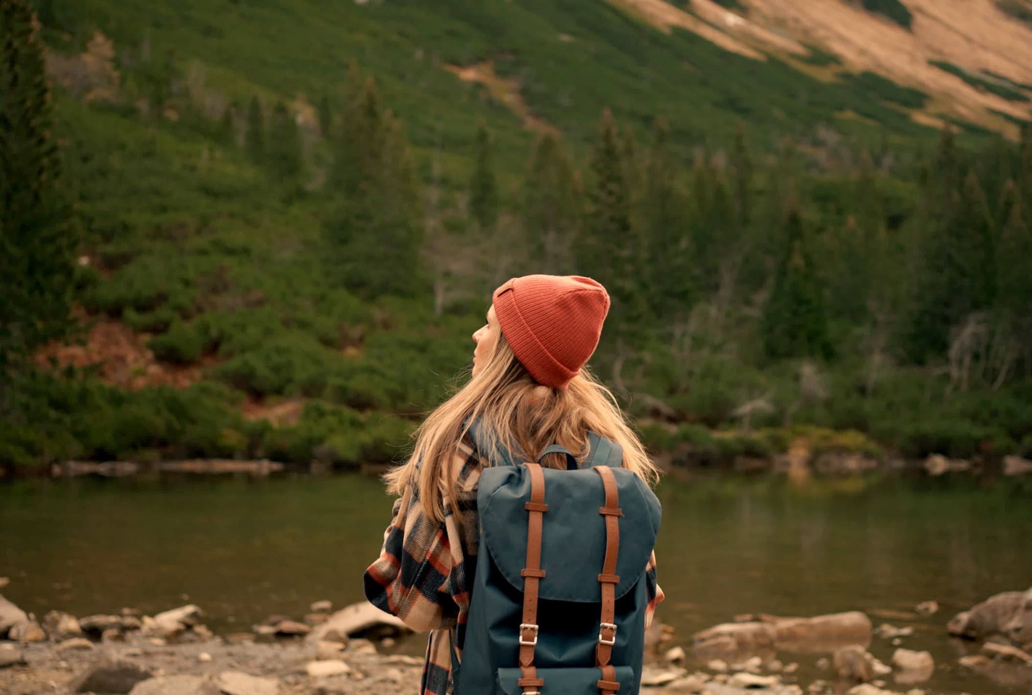 A hiker in a coral beanie and teal backpack standing at the edge of an alpine lake, turned away, looking across to a pine slope